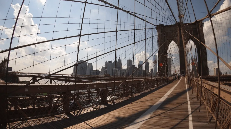 Explore NYC like a local by walking the iconic Brooklyn Bridge. Image taken from the central point of the bridge, surrounded by the wires that suspend it with New York City in the distance