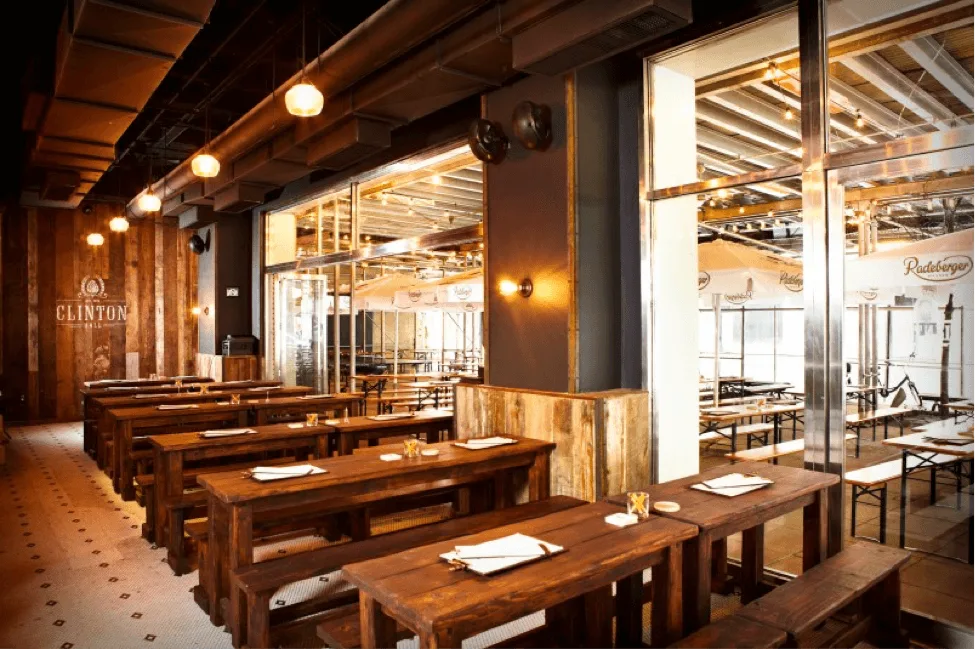 Photograph of the inside of an emplty restaurant with dark wooden tables and bench seats lining the side nearest, separated by a glass wall where there are lighter wooden tables and bench seats with metal legs