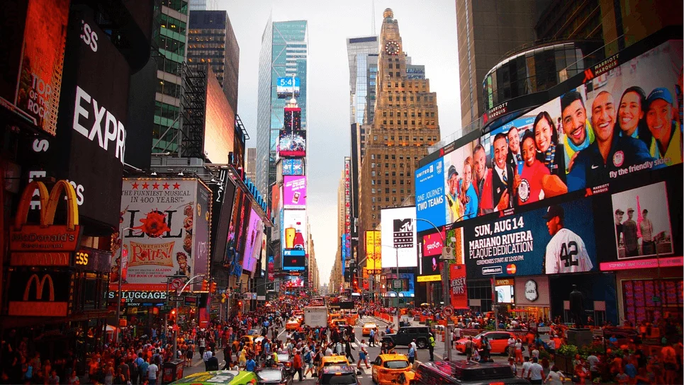 Elevated shot of Time Square in New York with neon advertisements either side of the street, masses of people walking in both directions and across the zebra crossing. In the foreground there are two new york taxi cabs waiting at the crossing. This vibrant scene makes it one of the best things to do in New York City.