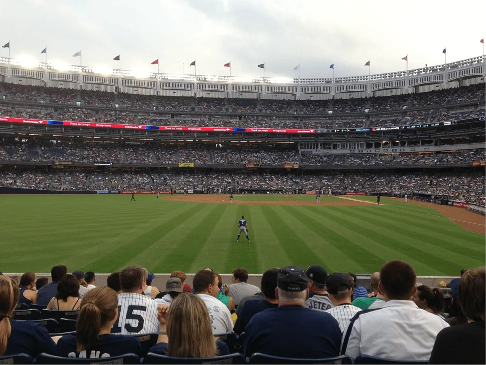 One of the best Things To Do in NYC is see a sporting event - this photo is of a Baseball Stadium from the lower stands with 7 rows of people sat in front, the ball game in the middle and full stands behind the game