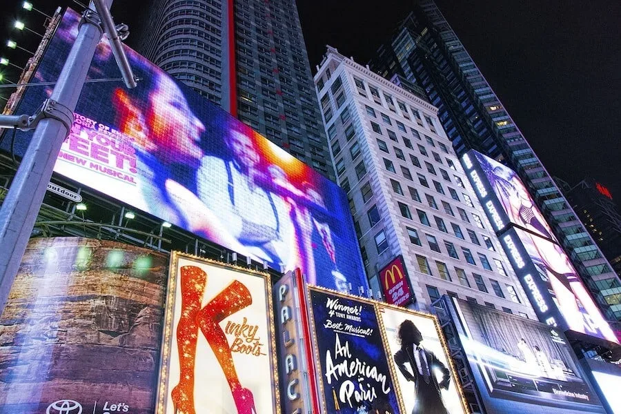 When wondering what to do in New York, seeing a show is a no brainer. Close up shot of Time Square from ground level looking up at the illuminated billboards advertising the Palace Theater, The Shows Kinky Boots and An American in Paris