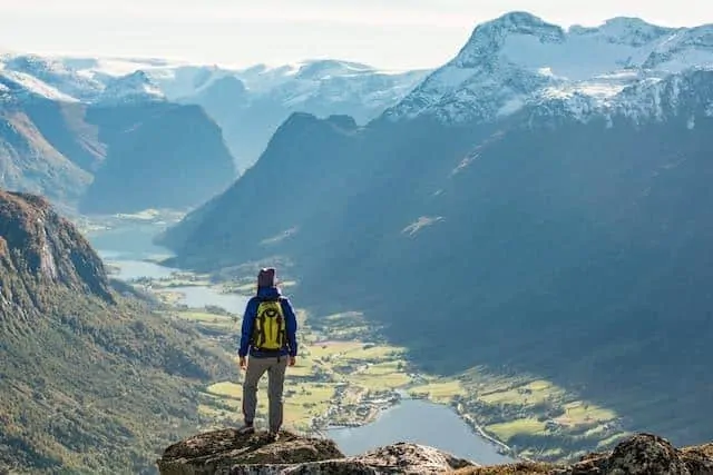 Man standing with his back to the camera at the top of a mountain overlooking a lush green valley with a lake in the middle