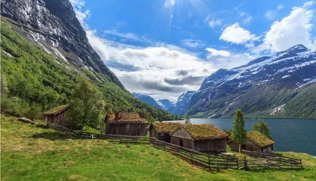 Loden Valley in Norway with 3 wooden houses in the foreground and lake, mountains and white clouds in the background