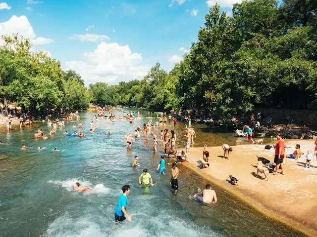 Busy outdoor swimming pool, lined with trees under a blue summer sky