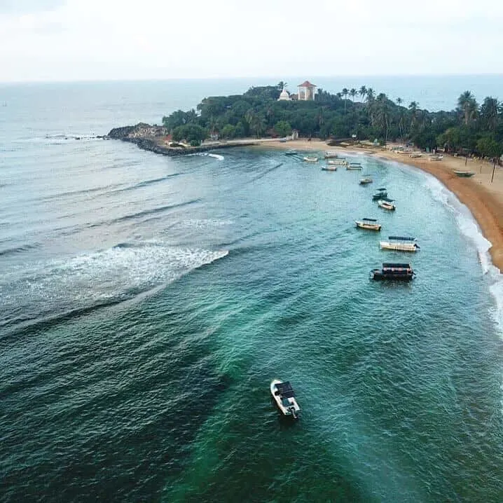 Unawatuna - beautiful beach in Sri Lanka, turquoise water in the foreground with boats lined up along the shore line, the beach curves round to the back of the image, behind which is a mount covered in plam trees among which you can see the white Pagoda and Big Buddha temple.