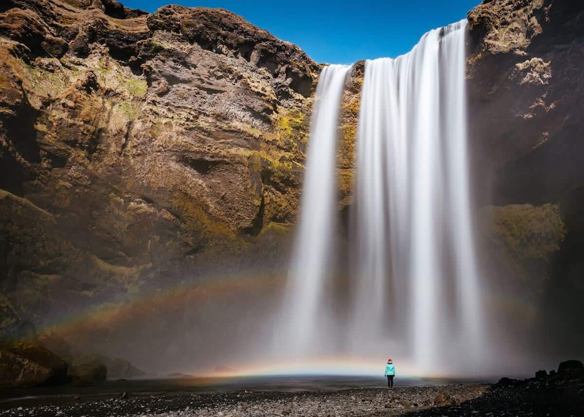 Cover Image for What to Pack for Iceland featuring a woman stood in a blue jacket and red hat in front of a waterfall in Iceland