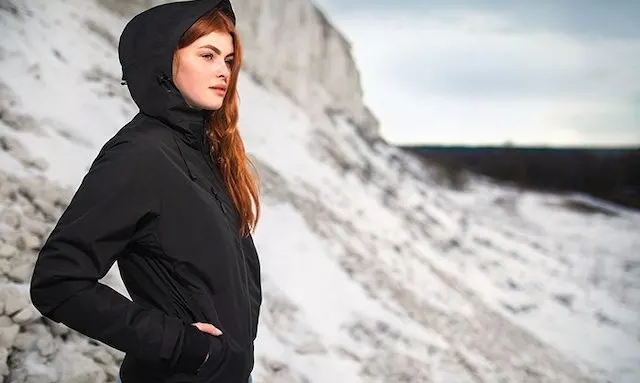 Red headed woman wearing a black jacket, her hands in the pockets, looking out in the distance in front of a rocky landscape