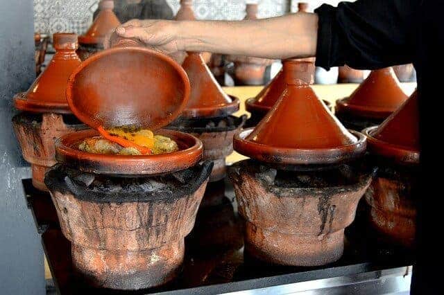 Tagines cooking on traditional heaters in Marrakech with a womans hand raising the lid on one to show vegetables cooking
