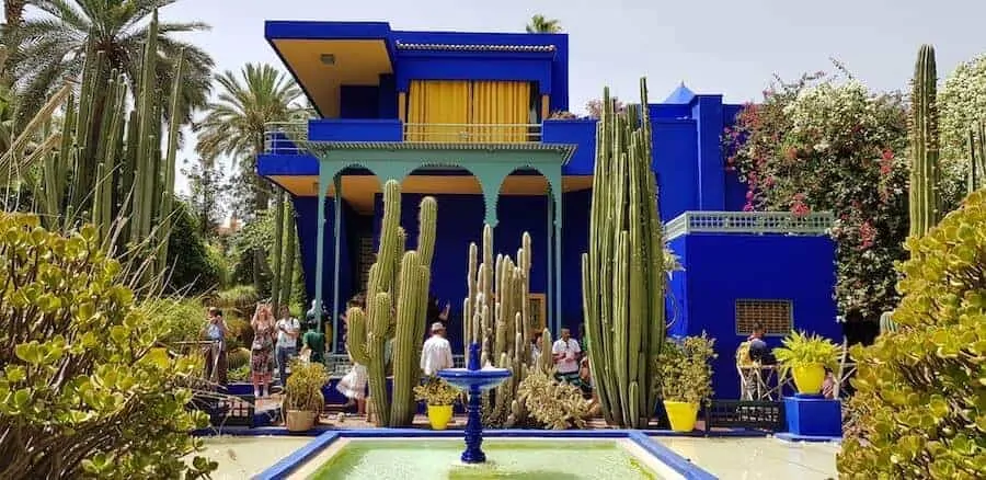 Striking blue building with contrasting yellow curtains in the upstairs windows and surrounded by exotic plants with a small blue fountain in the foreground