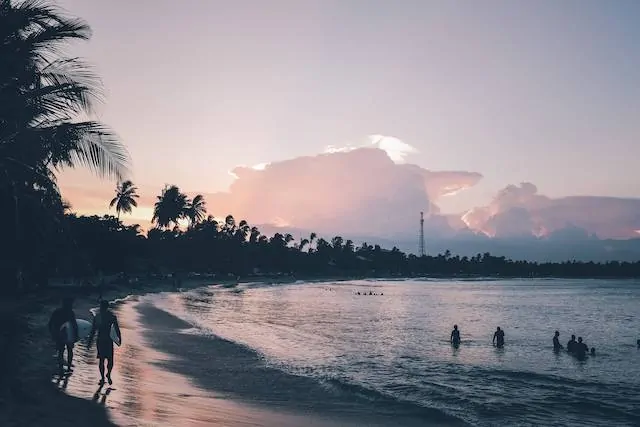 Dusk at Aragum Bay in Sri Lanka with purply pink skies, six people in shaddow in the ocean , two surfers with surf boards walking towards the camera and fluffy clouds in the sky