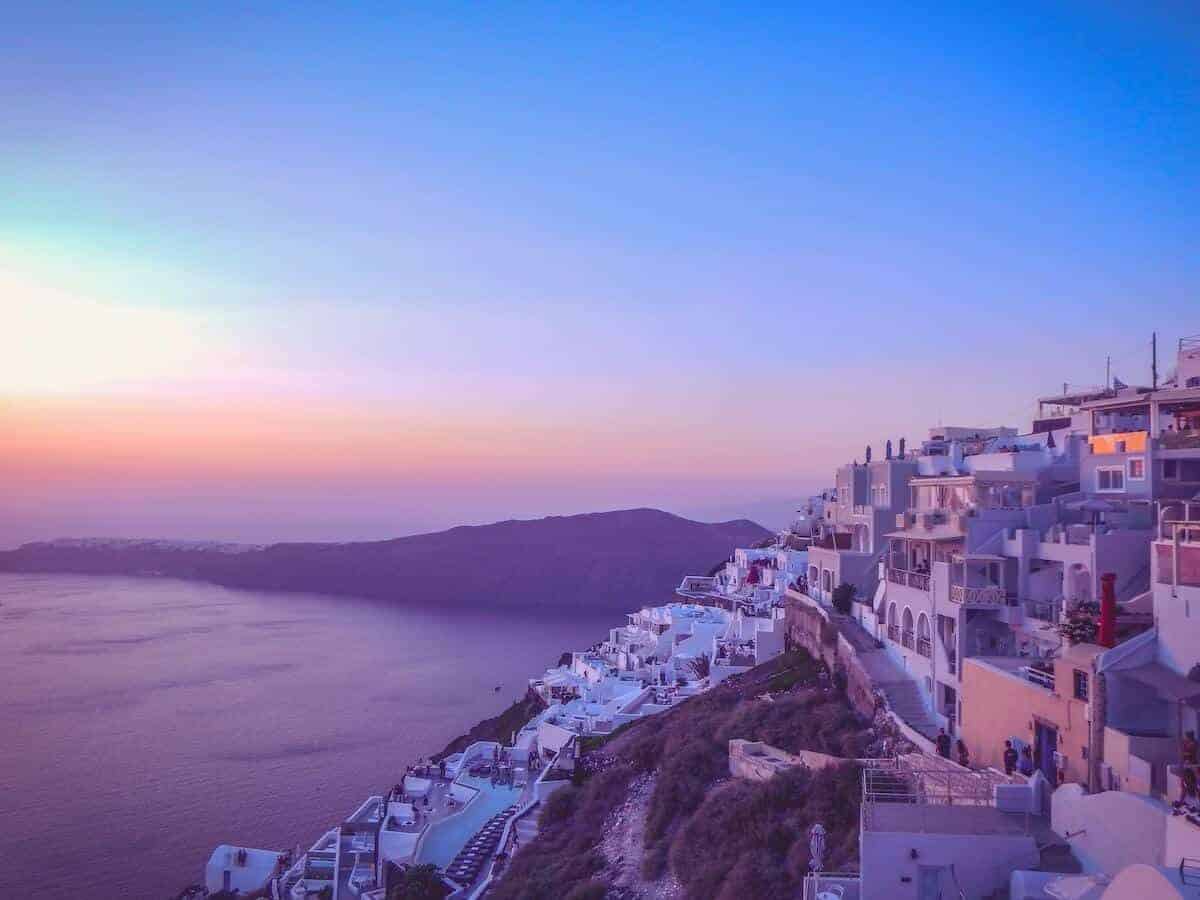 Cover photo for the Greek Island Hopping Itinerary and Travel Guide featuring a purple and pink sunset over a hillside village in Greece with the ocean below and mountains in the distance