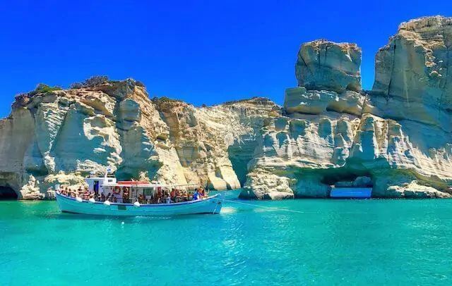 A small tourism boat packed with people sails on turquoise blue waters in front of a rugged rock face stretching the width of the shot in Milos