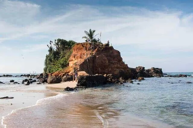 Mirissa Beach in Sri Lanka - prominent rocky outcrop in the centre of the frame with waves lapping around from each side. On the top of the large rock is a green plam tree, and the left side is completely covered with green climing plants