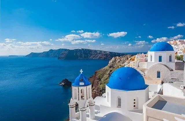 White buildings with the iconic blue dome site in the foreground of the image, set high on a hill. In the background the blue ocean and in the distance a neighbouring island rises out of the ocean