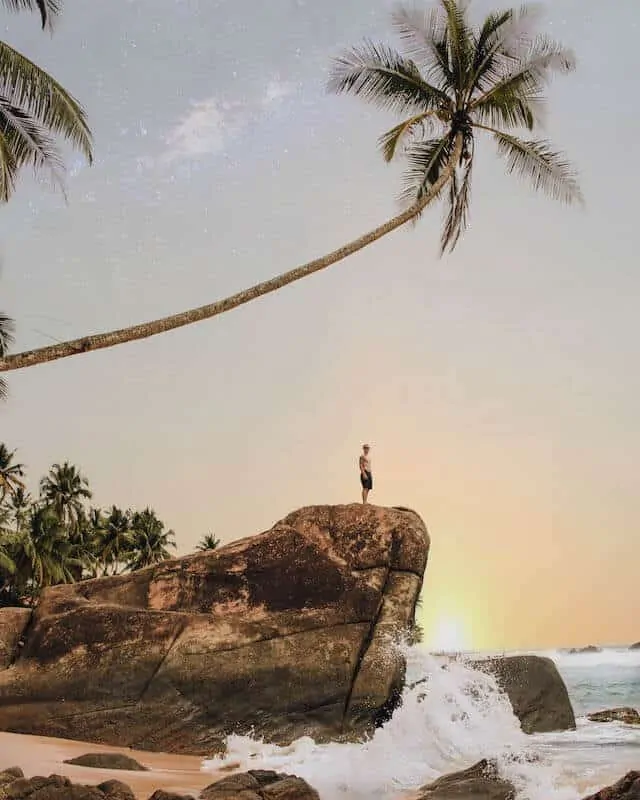 Image of Wijaya Beach - ocean on the right, on the left there is a large rock outcrop which has a singular palm tree hanging over on a 45 degree angle. A lone topless man with board shorts on is stood on the rock looking at the water