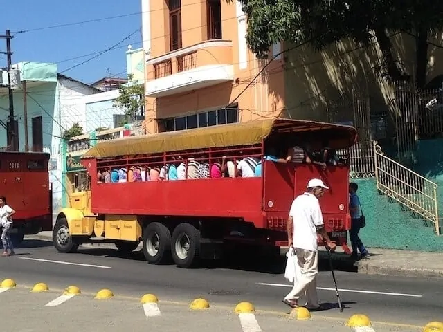 Red passenger trolley packed with people known as a Collectivo in Santaigo de Cuba parked in front of a light orange coloured building with a man dressed in white top and cream pants wearing a white baseball cap and using a walking cane to cross the road towards it
