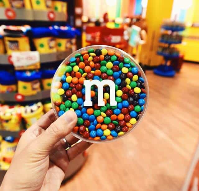 Hand holding a round clear plastic tub filled with multicoloured M&Ms in the M&Ms shop