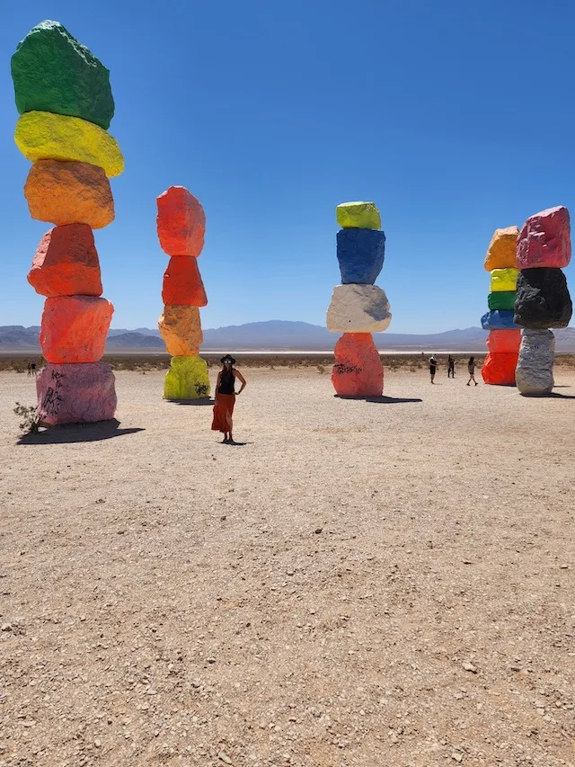 Woman in orange skirt, black top and black hat satnding in front of seven colourful rock stacks standing 20 feet tall