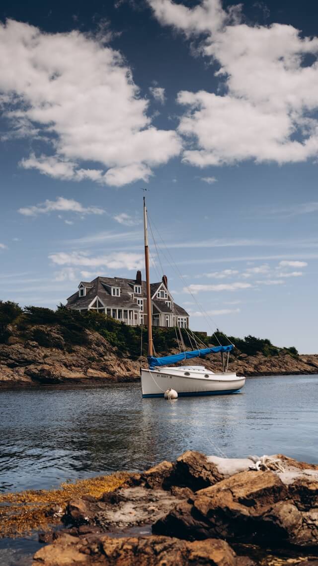 A large house on the side of a lake, a small white sailboat moored on the rocky shoreline in Newport Rhode Island