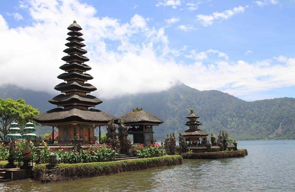 Tall brown wooden pagoda rising towards a blue sky with white clouds next to a lake - cover shot for the best things to do in Bali and guide to the best places to visit in Bali 