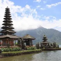 Tall brown wooden pagoda rising towards a blue sky with white clouds next to a lake - cover shot for the best things to do in Bali and guide to the best places to visit in Bali