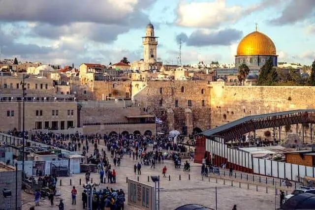 Western Wall surrounded by people, to the right is the walkway to the Temple Mount and Dome of the Rock sits on the raised platform at the back of the shot