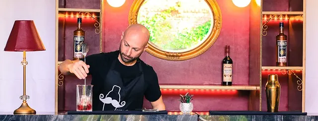 Bald Bartender wearing black shirt with cocktail shaer motif stiring a cocktail on the bar in front of him