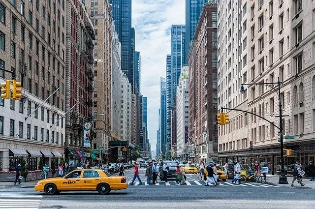 New York City street with high rise buildings either side, a yellow cab in the foreground and people walking aross a zebra crossing spanning the width of the street