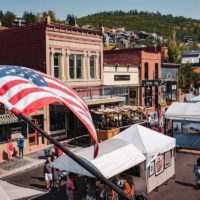 Cover photo from Things to do in Park City Utah featuring an aerial view of main street in Park City in summer with an American flag in the foreground and white tents of the market lining the street