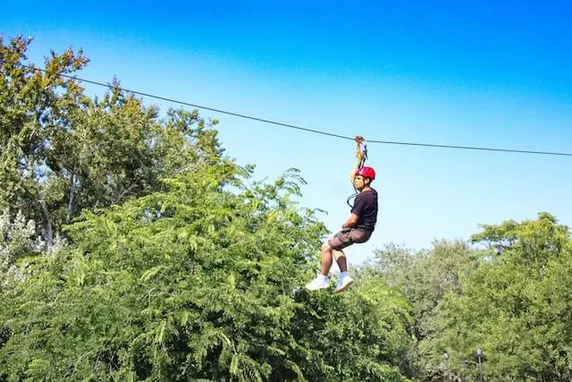 Man wearing a red crash helmet hanging from a zipline above a line of treetops