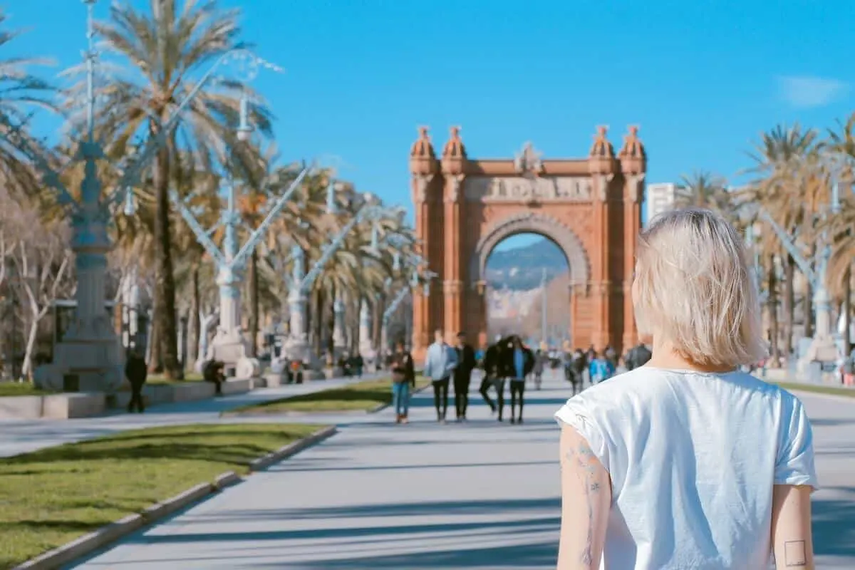 Barcelonas Red Moorish Style Arc de Triomf at the end of a grey concrete pathway through a park; in the forground a woman with blond hair and a blue shirt has her back towards the camera looking towards it. This is the header image for the Best Places to Visit in Barcelona