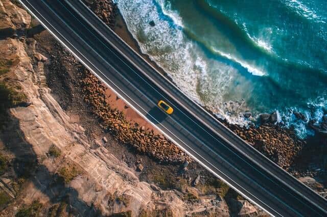 Aerial shot of a road with cliffs on one side and ocean on the above, taken directly above a bright yellow open top car