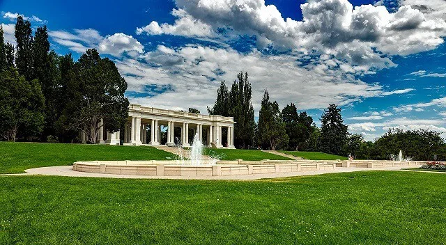 White Columade greco-roman style structure surrounded stands behind a simple fountain surrounded by green grass and under a bright blue sky 90% covered in light white clouds in Cheesman Park Colorado