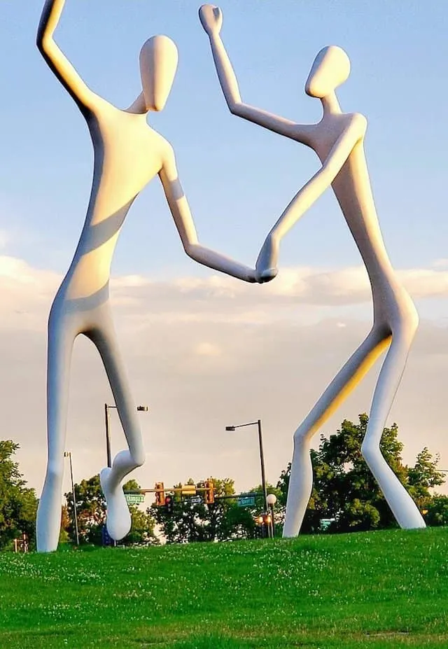 Two 60 foot high white fiberglass sculptures holding hands and dancing on a lawn