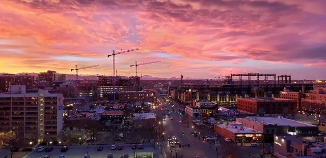Aerial shot of Downtown Denver with several cranes rising above the buidlings and consrtuction sites at dusk with lights on in the buildings and on cars underneath a cotton candy pink sunset sky