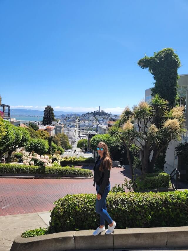 Woman wearing blue jeans, white tennis shoes and a black leather jacket with mirrored sunglesses standing on Lombard Street in San Francisco