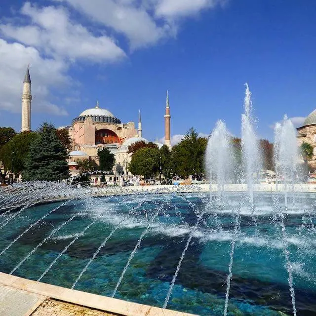 View of Sultanahmet Square in Istanbul taken from behind a fountain with the Pink Facade of the Hagia Sofia flanked by two minaret spires in the distance