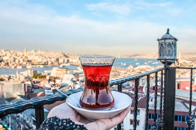 Woman holding a white plate with a tulip glass of Turkish Tea on a rooftop overlooking the Bosphoros river with the city of Istanbul in the background