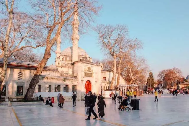 Open square dotted with people in front of the white facade of the Suleymaniye mosque domed structure with 2 minaretes standing either side