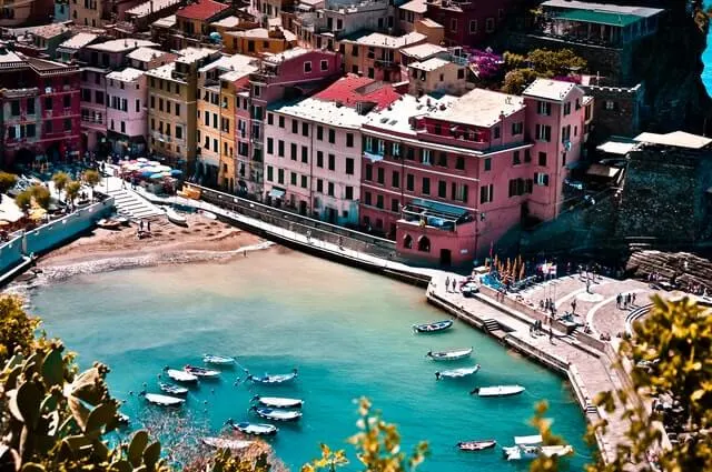 Aerial view of a harbor in Cinque Terre Italy with small white boats flating on clear blue waters with pink, white and orange fronted buildings along the wall of the harbor 