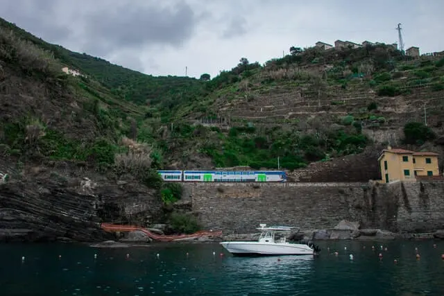 White and Green Train moving along tracks elevated on a stone wall above the ocean. There is a white boat floating in the foreground with green hills in the background.