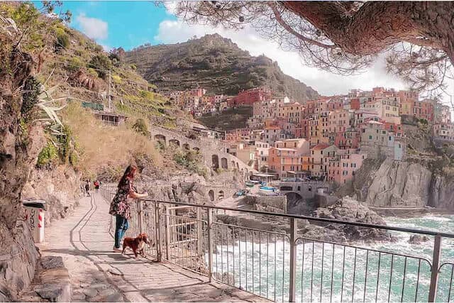 Elevated walkway heading towards a village in Cinque Terre with the ocean on the right and a woman with a dog on a lead standing at the rails along the path looking out over the ocean
