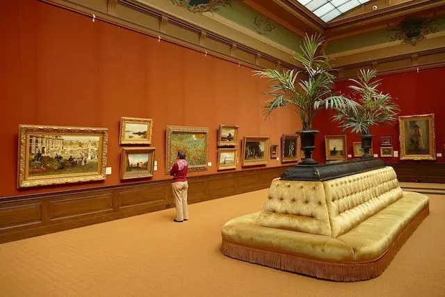 Inside Tylers Museum with white leather central seat, wooden walls with art work hung at eye level, a woman admiring the art wears a red sweatshirt