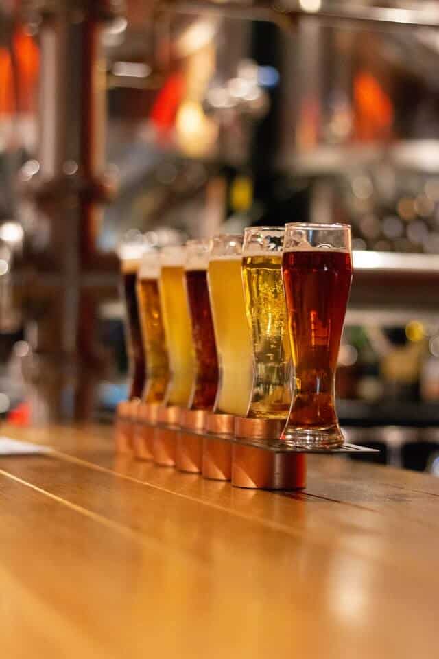 Flight of different types of beer in tall glasses on a wooden stand on top of a bar