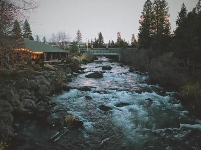 Fast flowing river with a restaurant building on the banks on the right and a bridge over the water in the distance