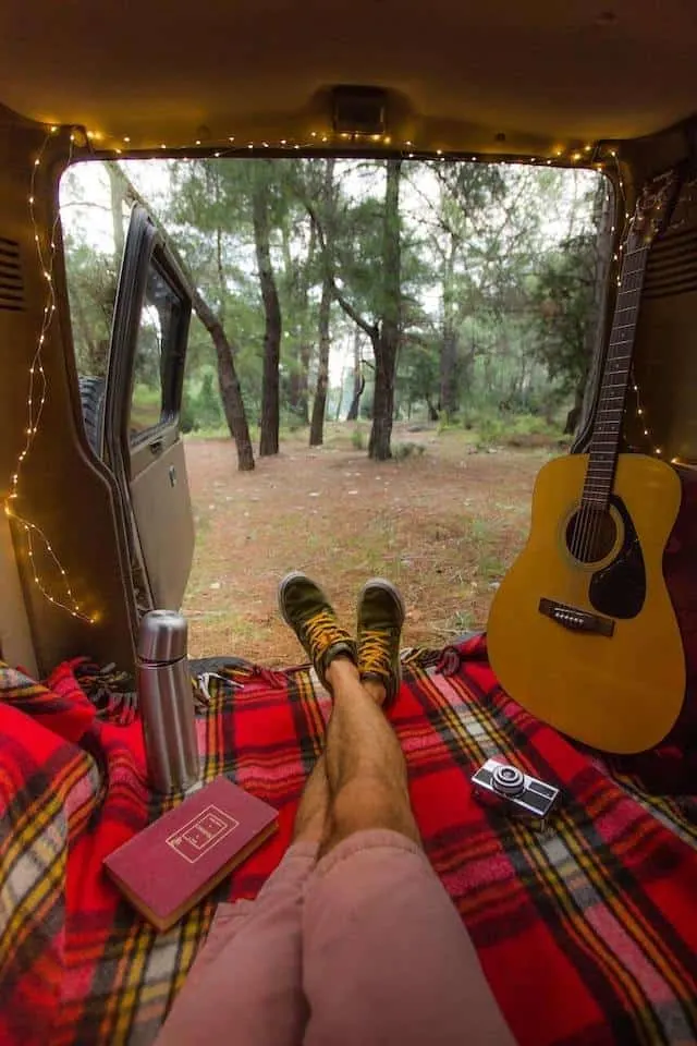 Campervan with the back doors open looking out into the forest. Mans legs wearing pink shorts pointed towards the open doors with a guitar sitting against the side of the vehicle on the right and the door framed by fairy lights