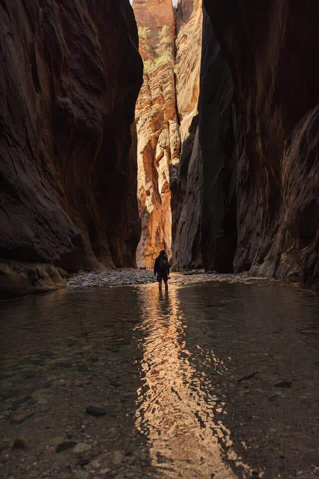 Man wading through narrow water between the high sides of the rocks which form the Narrows hiking trail in Zion National Park