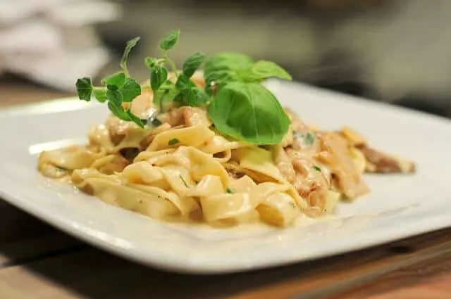 White plate containing wide ribbons of pasta in a white suace with mushrooms topped with a sprig of green leafy basil