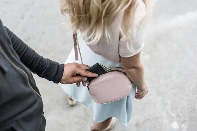 Mans hand taking a purse from a womans pink bag worn across her body and behind her back. She has blonde hair, a white shirt and a light blue skirt