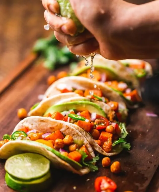5 Tacos on a wooden board lined up side by side, an hand hovering above them squeezing lime on to them